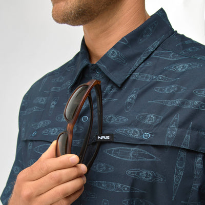 Close-up of man wearing navy NRS Men's Gear Shirt with kayak pattern, holding sunglasses clipped to chest pocket.